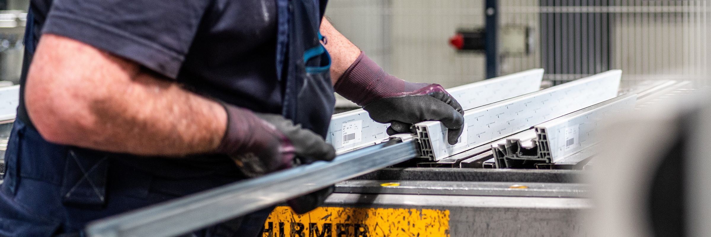Hände in Arbeitshandschuhen führen ein Fensterprofil in eine Maschine.
