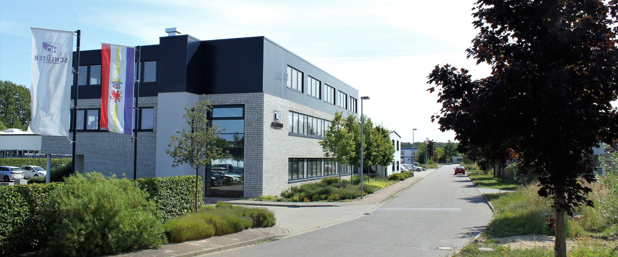 Das Bürogebäude von H.O. Schlüter am Gewerbering 6 in Lübz, davor Fahnen mit Firmenlogo und der Landesflagge Mecklenburg-Vorpommerns.