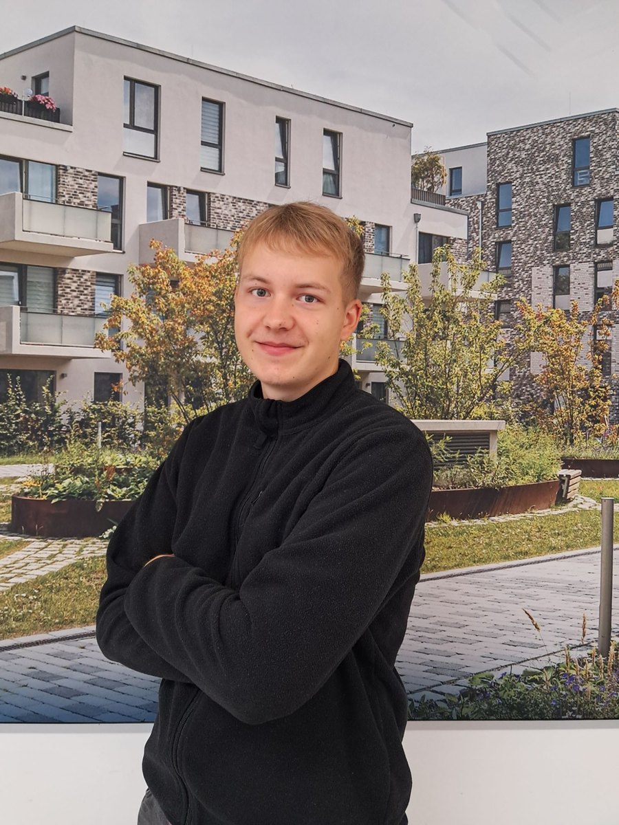 Felix Schlapmann mit verschränkten Armen vor einem Bild mit Wohngebäuden.