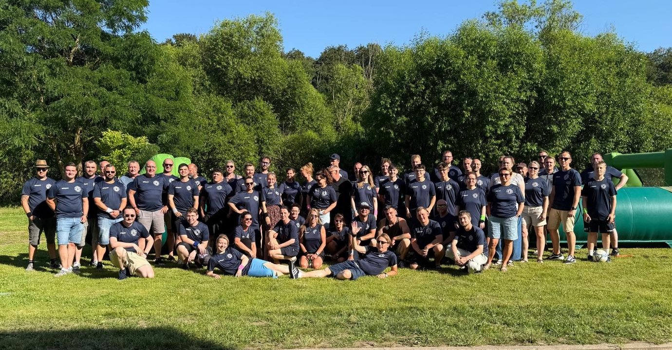 Die Belegschaft in dunkelblauen Team-Shirts beim Gruppenfoto auf einer Wiese, im Hintergrund Bäume.